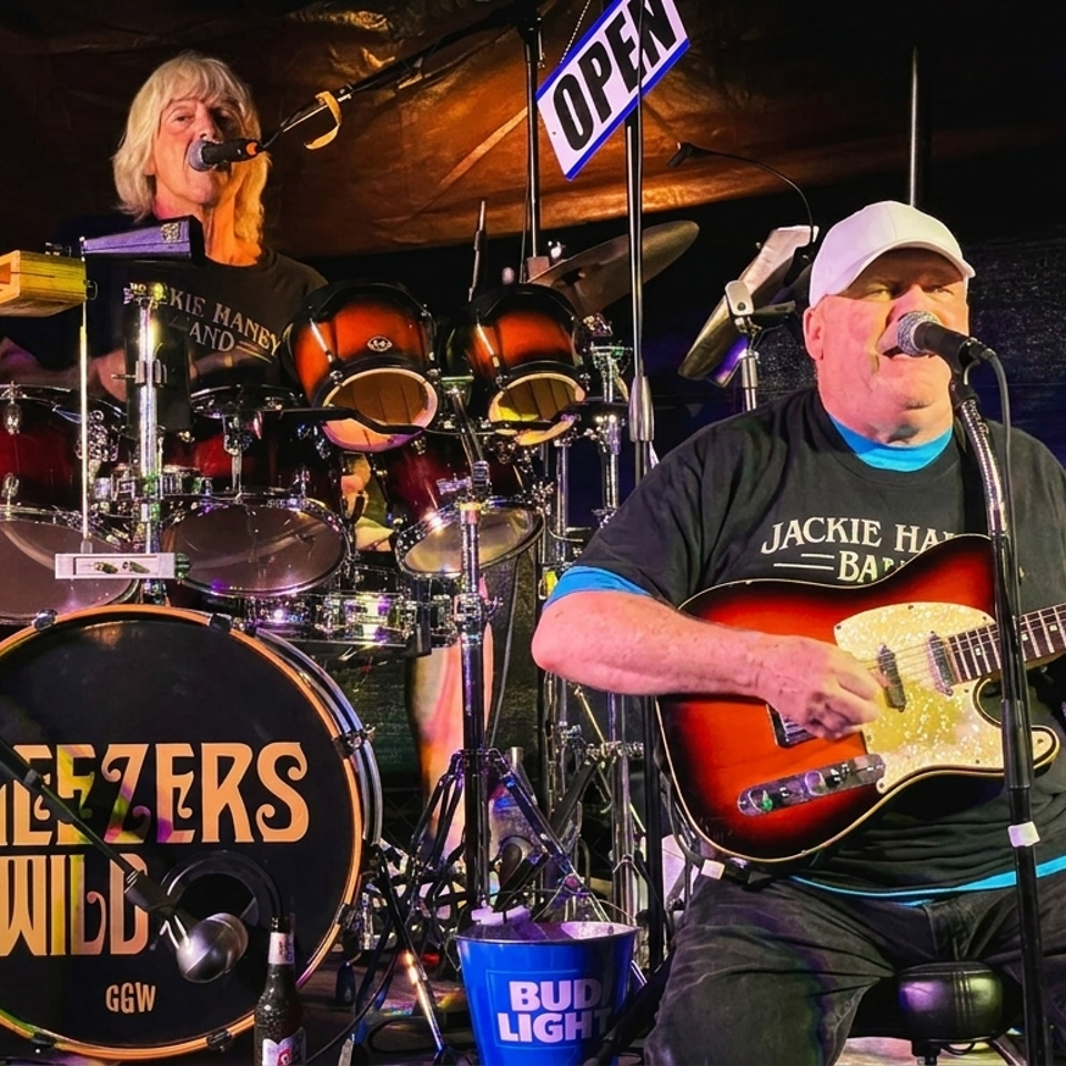 Two musicians perform onstage, with a guitarist singing beside a drummer, drum kit, amplifiers, and an open sign.